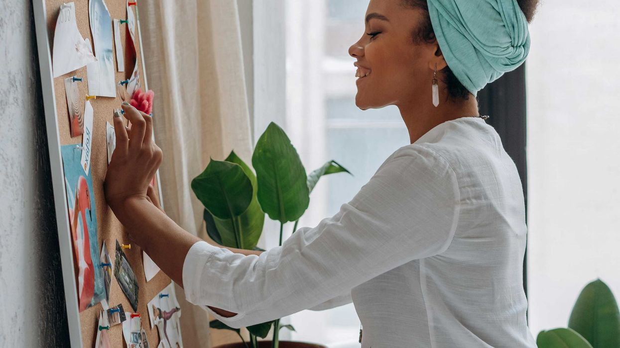 Woman pinning paper on a board, smiling in a bright room with plants.