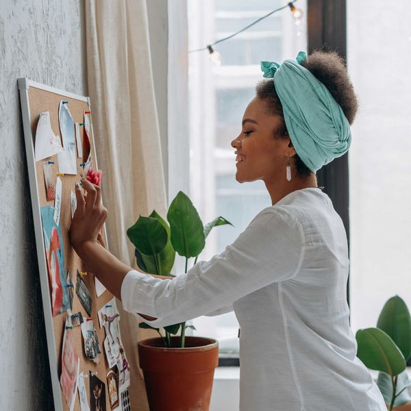 Woman pinning paper on a board, smiling in a bright room with plants.