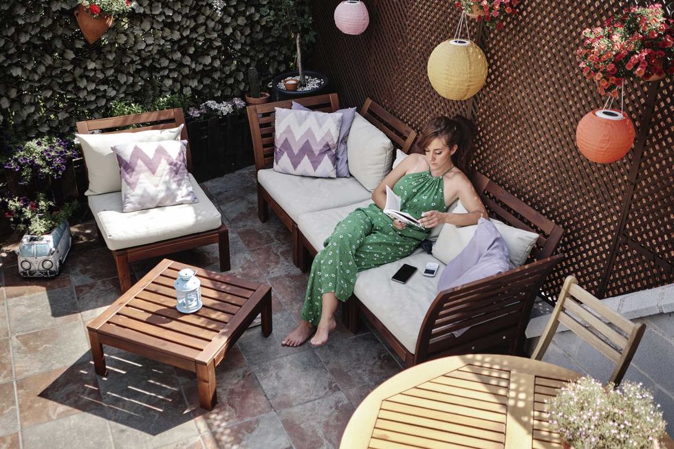 Woman reading on patio sofa surrounded by plants and colorful lanterns.