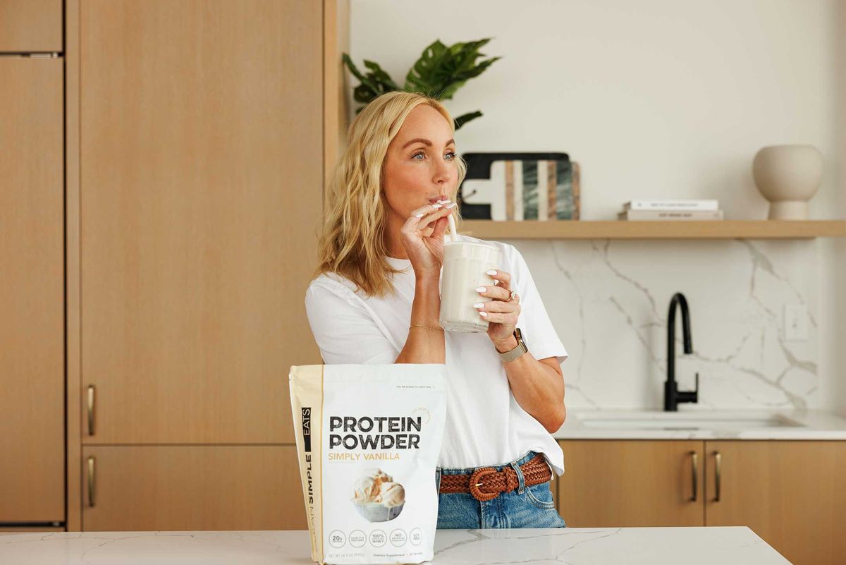 Woman sipping protein shake in kitchen with protein powder bag on counter.