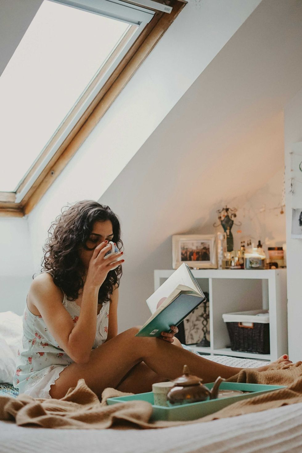 Woman sipping tea and reading a book by a skylight window in a cozy bedroom.