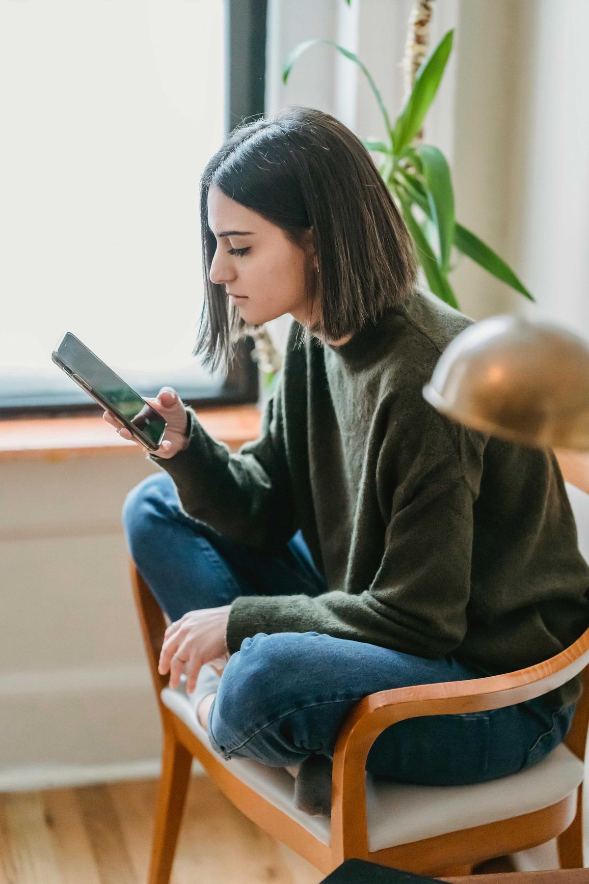 Woman sitting in a chair, focused on her smartphone, with a plant in the background.