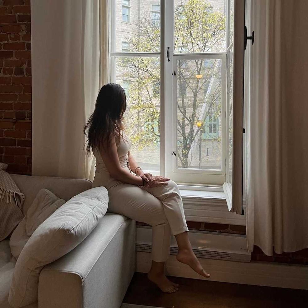 Woman sitting on sofa, gazing out window at tree-lined street.