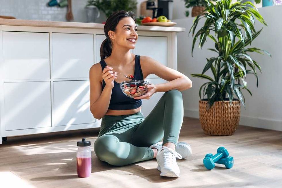 Woman sitting on the floor, eating a salad with dumbbells and drink nearby.