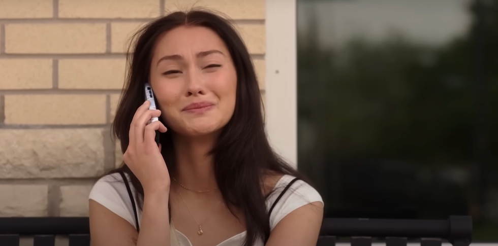 Woman sitting, talking on phone, with a bittersweet smile, against a brick wall.