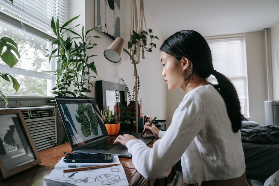 Woman sketching on a laptop at home, surrounded by plants and art supplies.