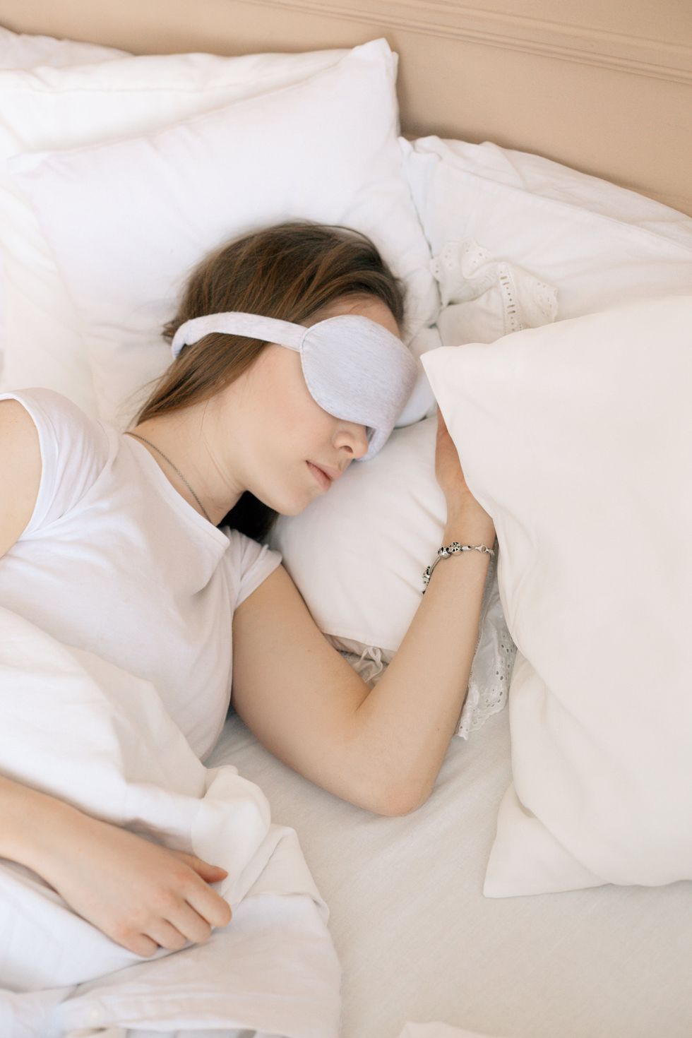 Woman sleeping with a gray eye mask in a bed with white sheets.