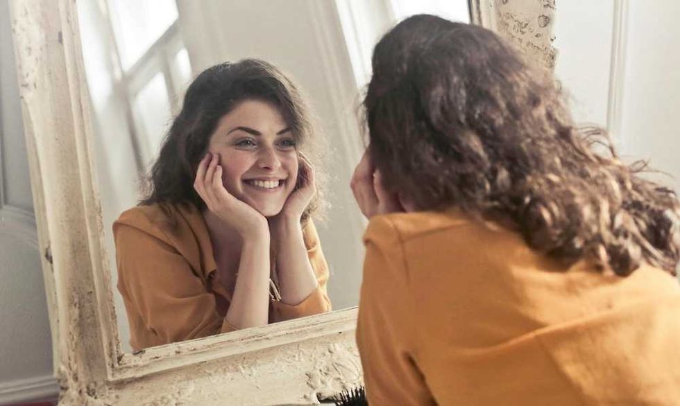 Woman smiling at herself in a mirror, wearing a yellow shirt, with natural lighting.
