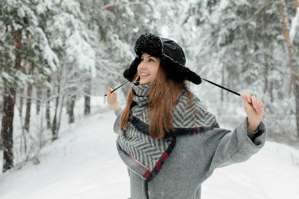 Woman smiling in snowy forest, wearing a winter coat and hat with ear flaps.