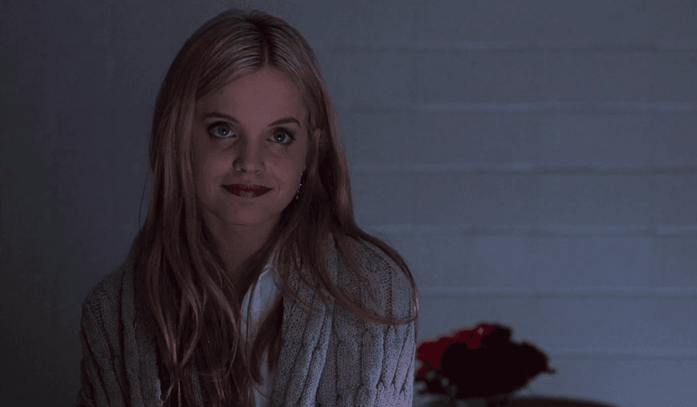 Woman smiling softly in a dimly lit room with flowers in the background.