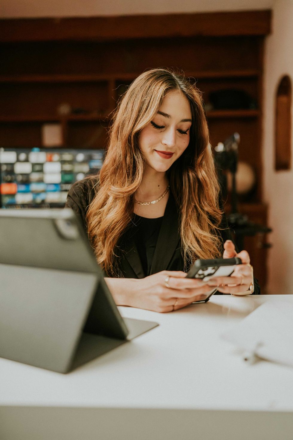 Woman smiling while using a smartphone at a desk with a tablet.