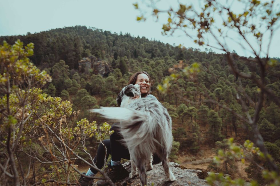 Woman smiling with her dog on a rocky hill, forested mountains in the background.