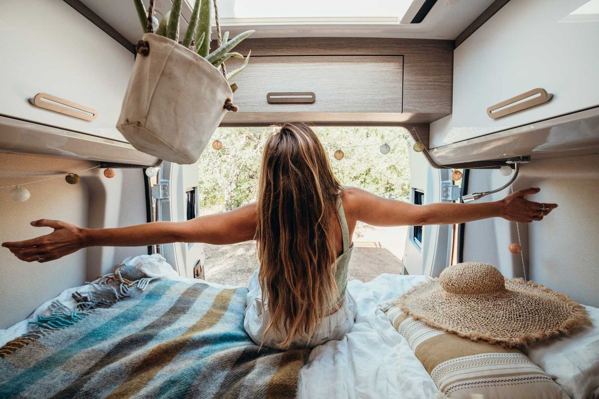 Woman stretching arms in a camper van, overlooking nature through open doors.