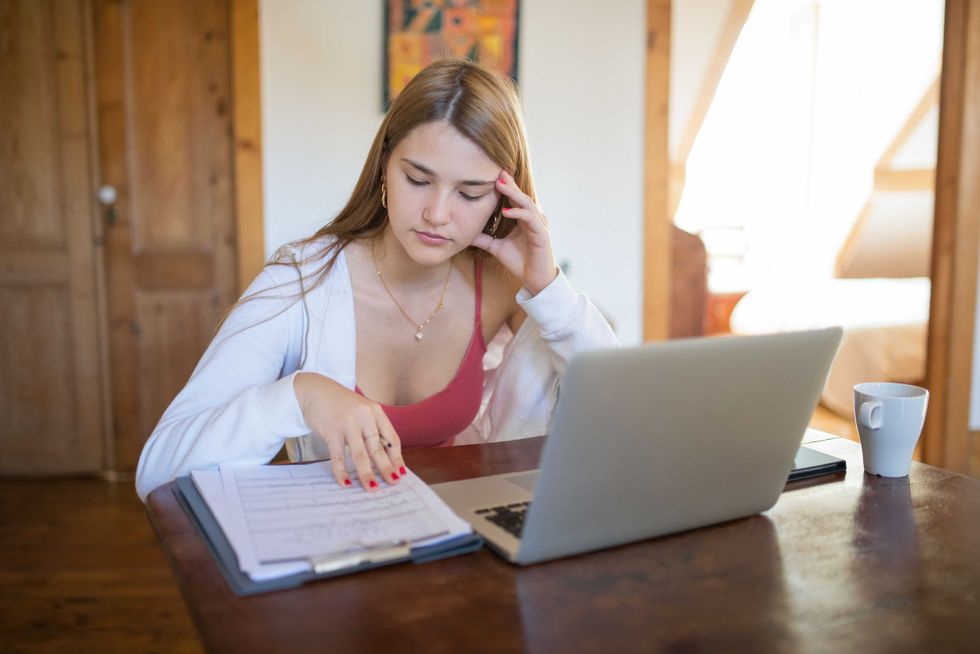 Woman studying at a desk with a laptop, clipboard, and mug.