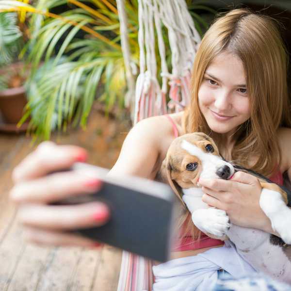 Woman taking a selfie with a dog while sitting in a hammock indoors.