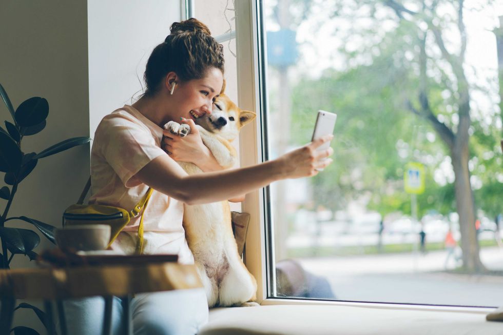 Woman taking a selfie with her dog by a window, wearing headphones and smiling.