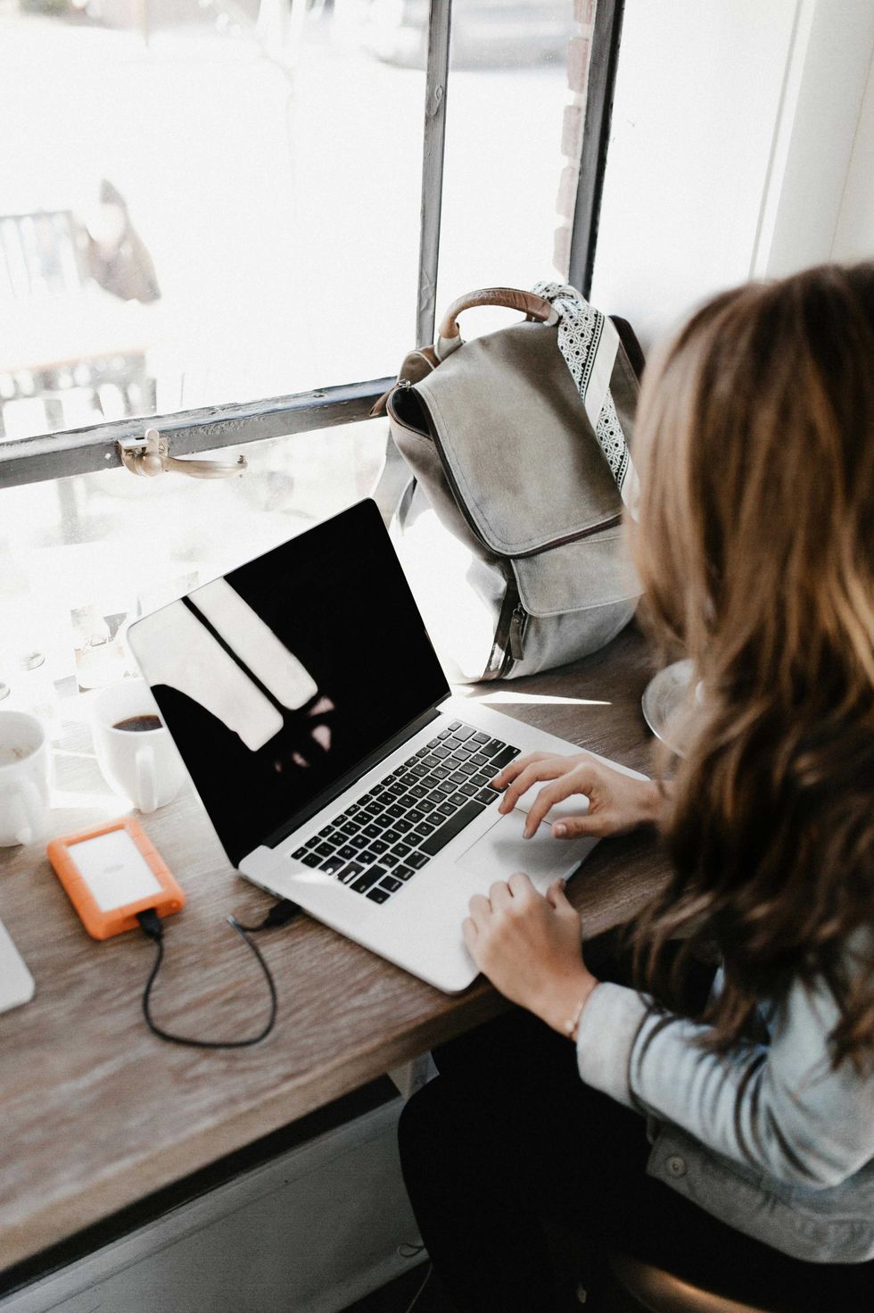 Woman using a laptop by a window with a backpack and external drive.