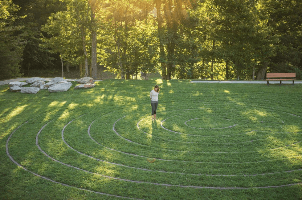 Woman walking in a grassy spiral labyrinth, surrounded by trees in sunlight.
