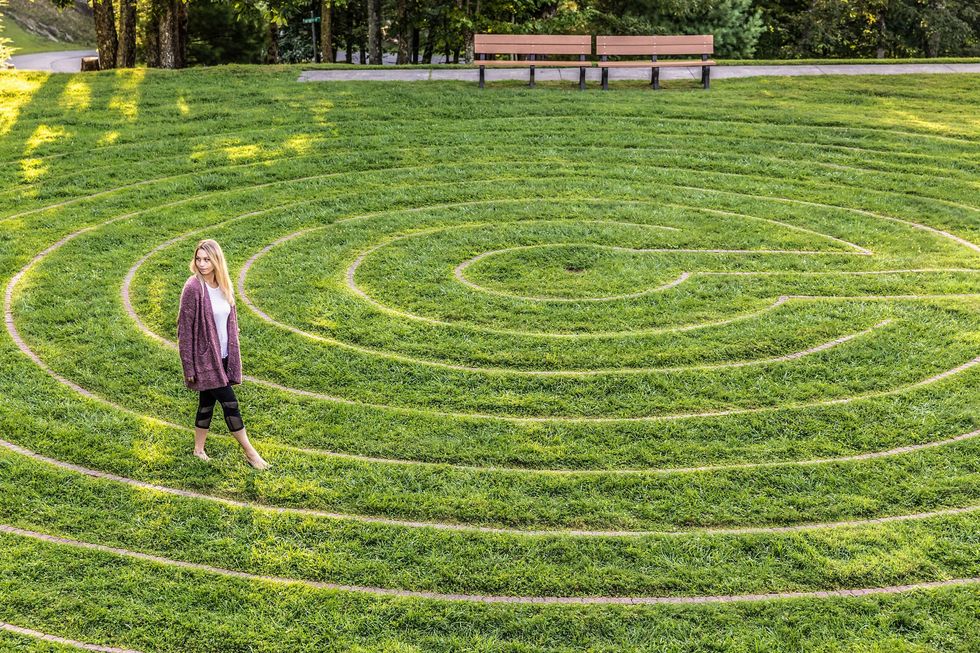 Woman walking on spiral path in grassy labyrinth with benches in background.