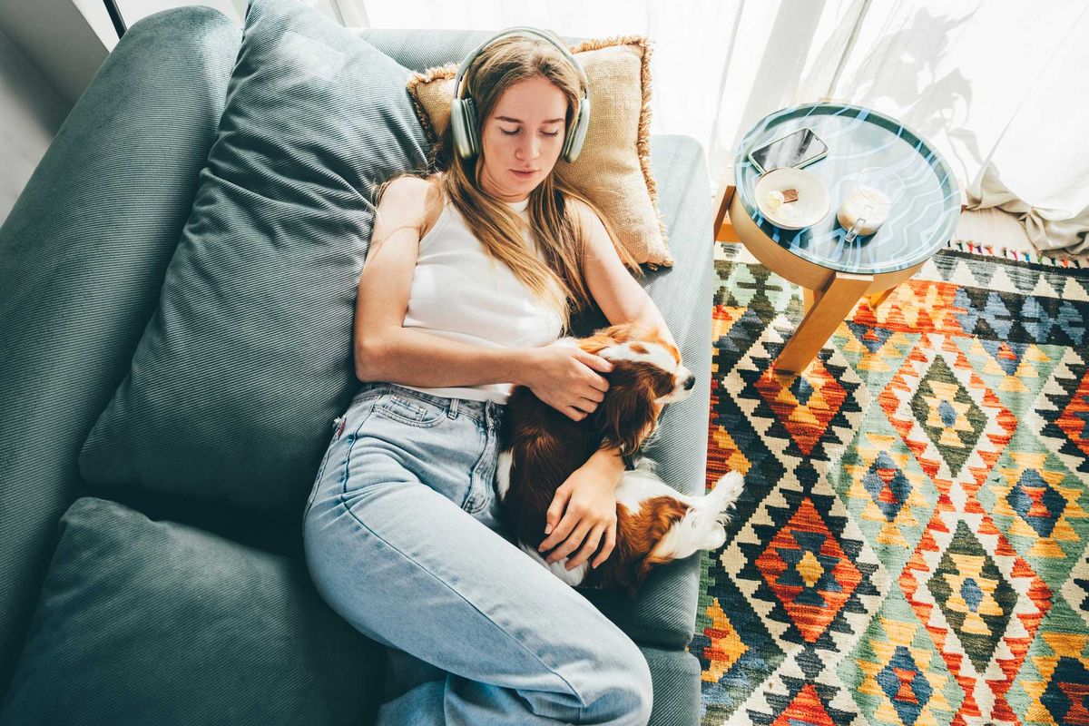 Woman wearing headphones, relaxing on sofa with dog, colorful rug nearby.
