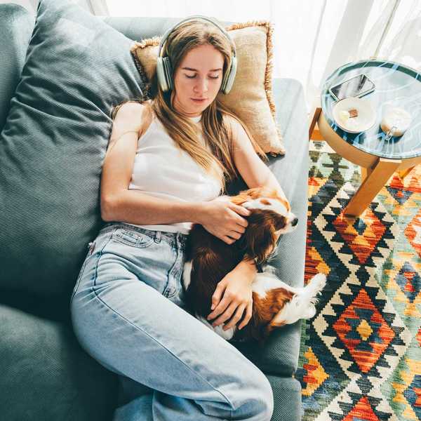 Woman wearing headphones, relaxing on sofa with dog, colorful rug nearby.