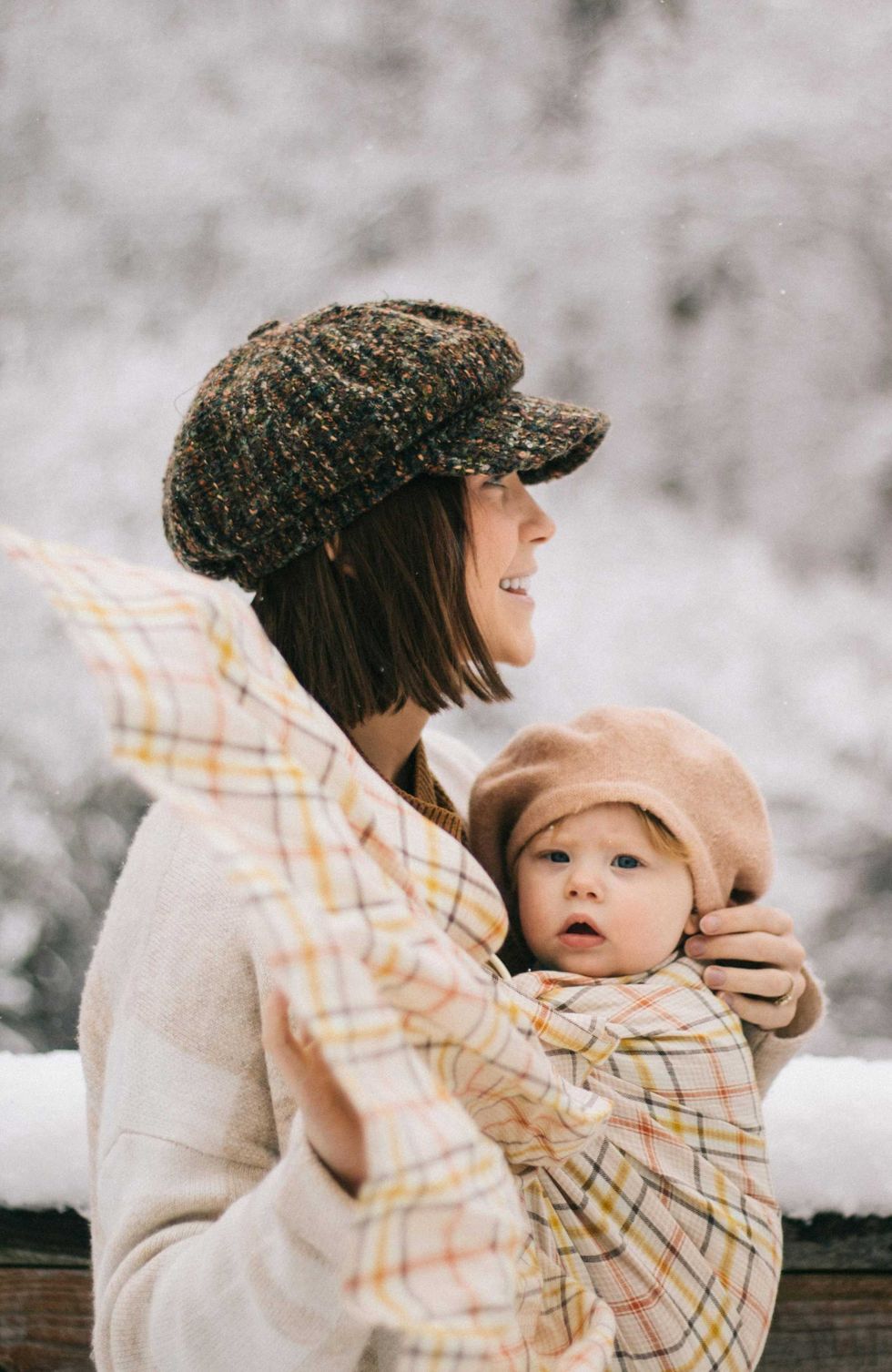 Woman with a baby wrapped in plaid, both wearing hats in a snowy setting.