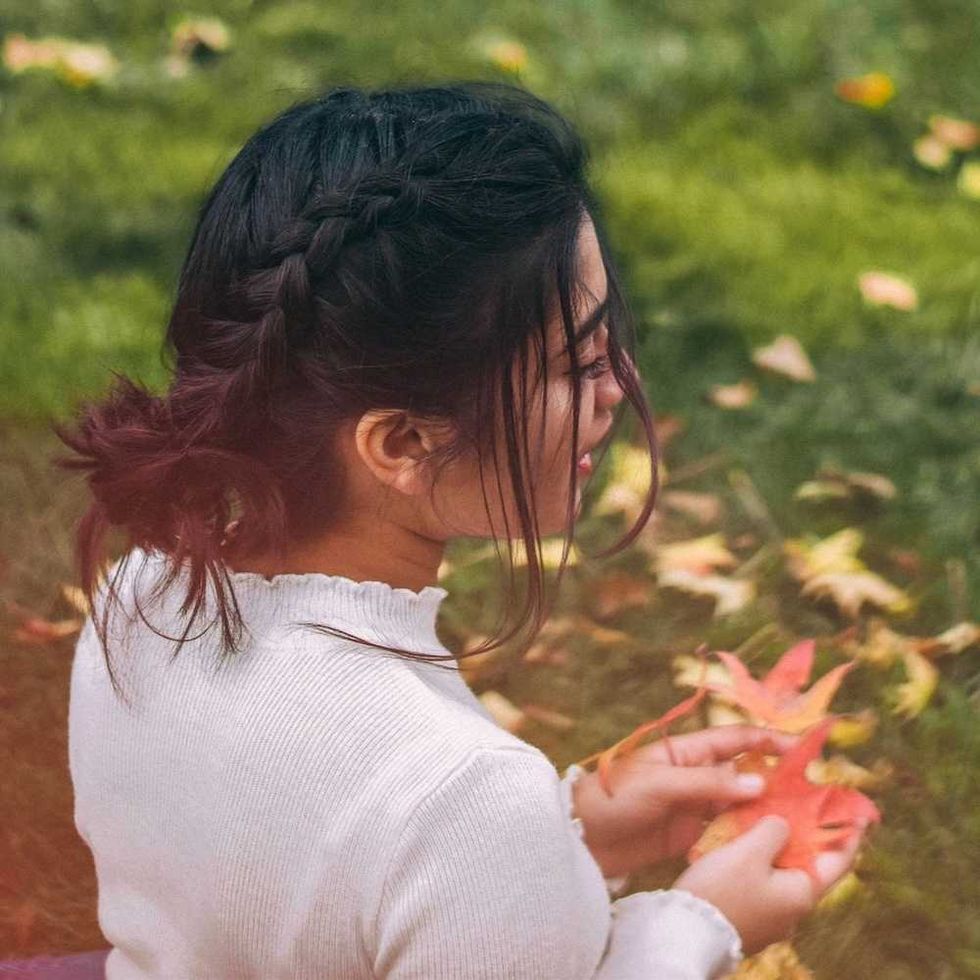 Woman with braided hair holds autumn leaves on a grassy field.