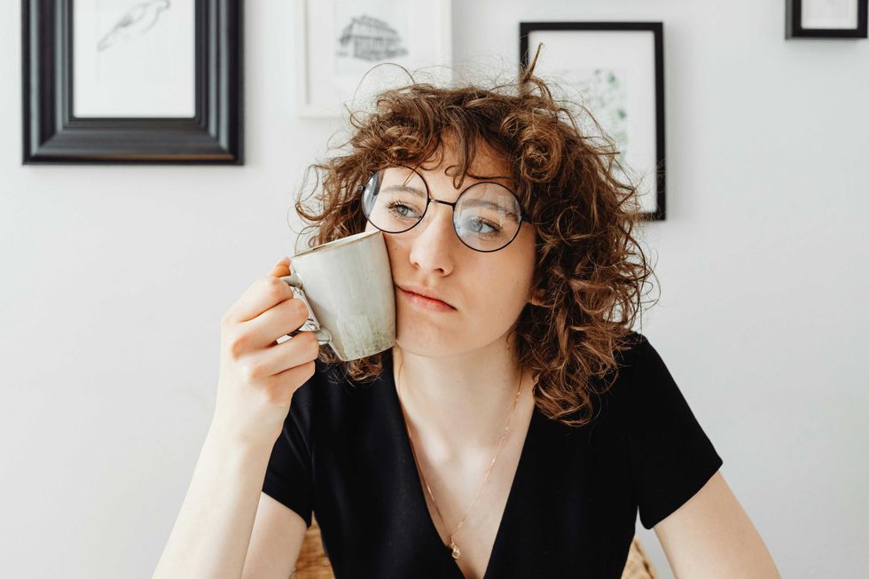 Woman with curly hair and glasses holding a mug, sitting thoughtfully indoors.