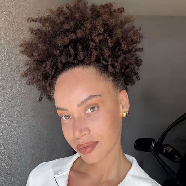 Woman with curly hair and gold earrings smiles in front of a gray wall.