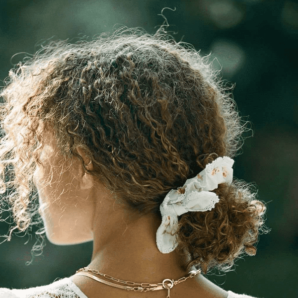 Woman with curly hair in bun, lace top, and gold necklace, with floral hair tie.