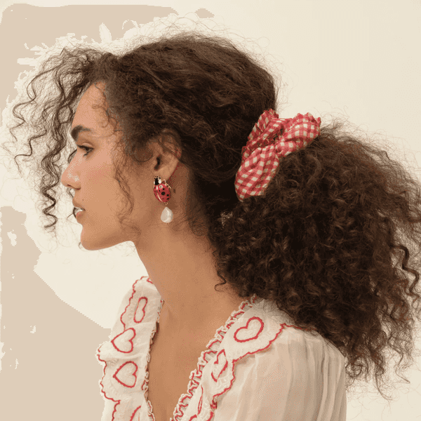 Woman with curly hair, red checkered scrunchie, heart-trimmed blouse, and ladybug earring.