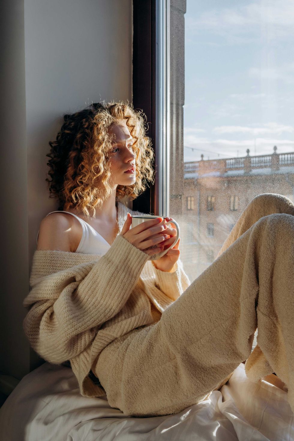 Woman with curly hair, wearing cozy clothes, holds mug by a sunny window.
