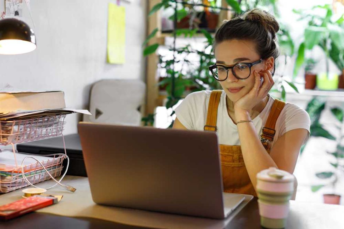 Woman with glasses working on a laptop, surrounded by plants.