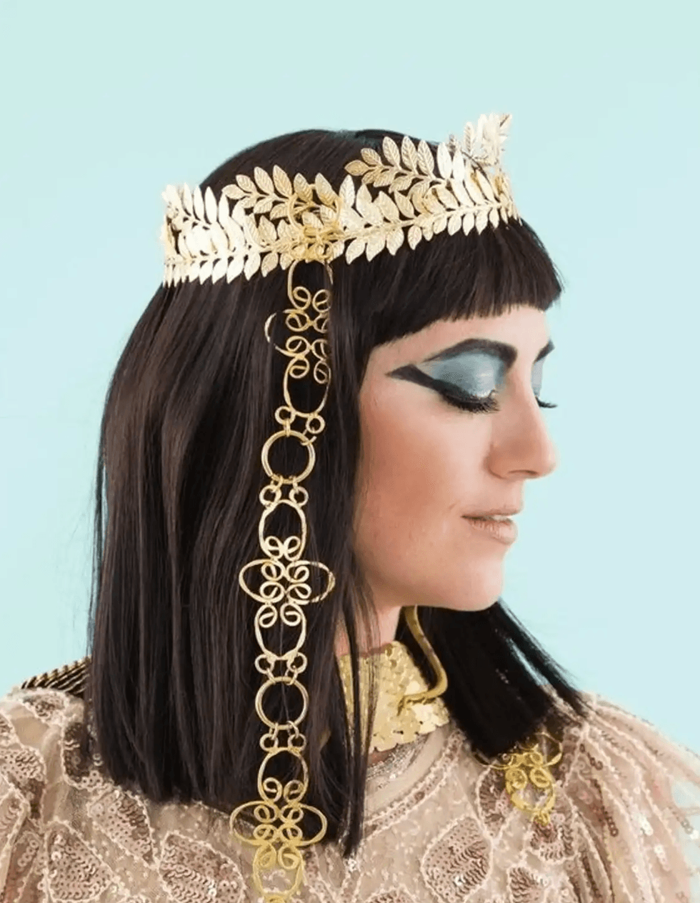Woman with gold laurel crown and ornate jewelry against a light blue background.