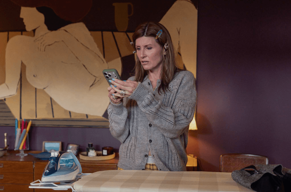 Woman with hair clips looks at phone, standing by ironing board.