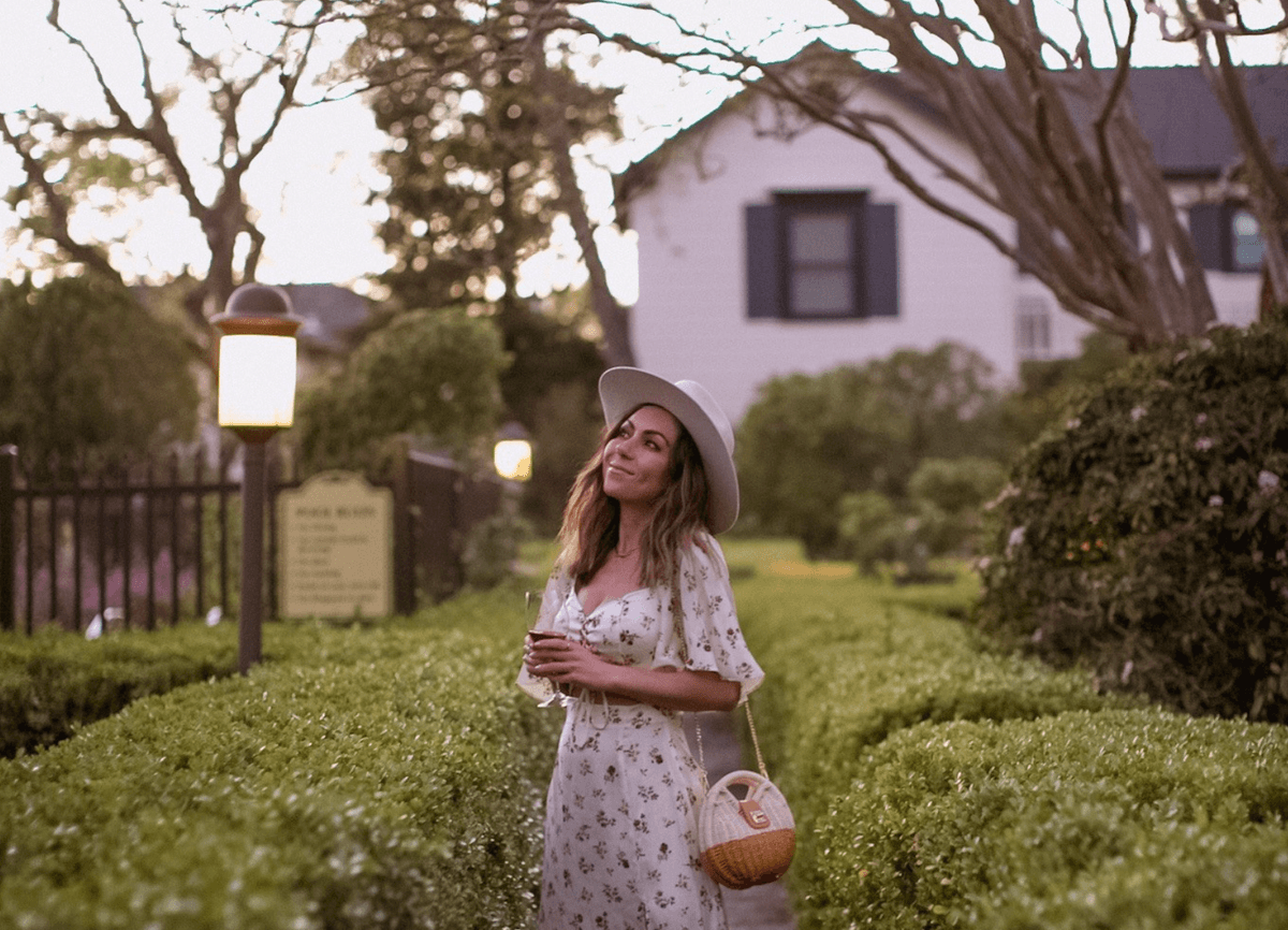 Woman with hat standing in a garden, holding a drink, near a lamp post at dusk.