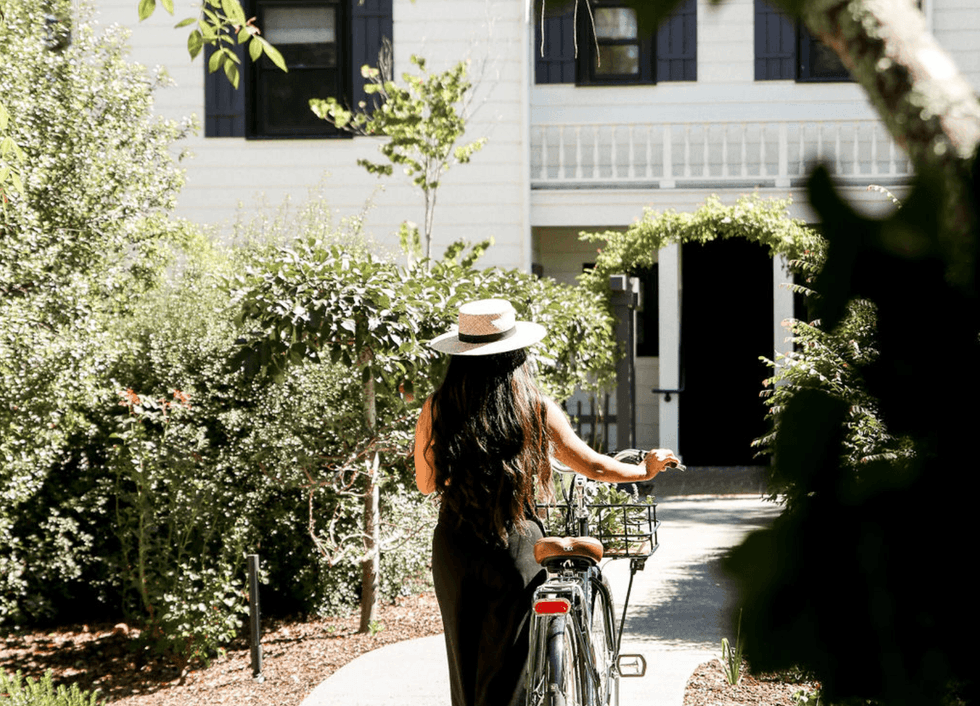 Woman with hat walking bicycle on a garden path towards a house.