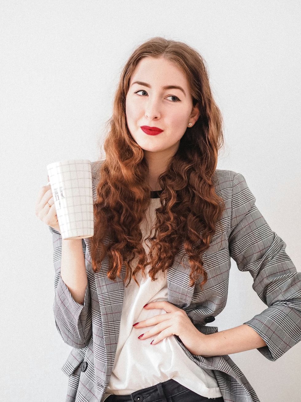 Woman with long curly hair holds a checkered mug, wearing plaid blazer against white background.