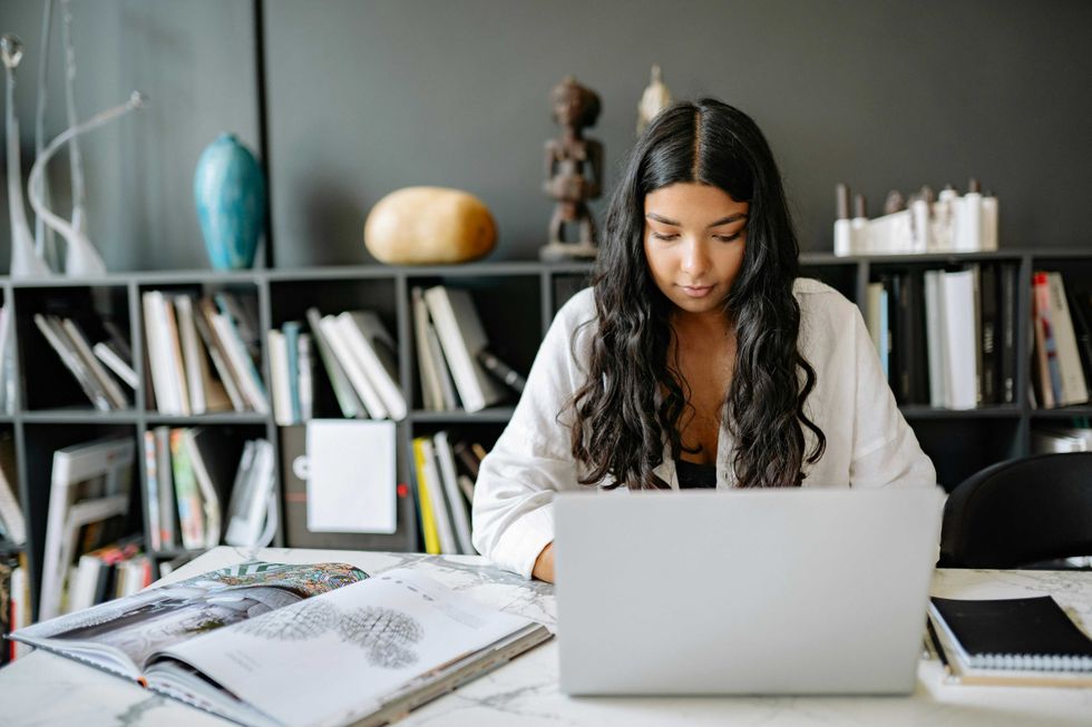 Woman with long hair working on a laptop at a desk with books and shelves in the background.