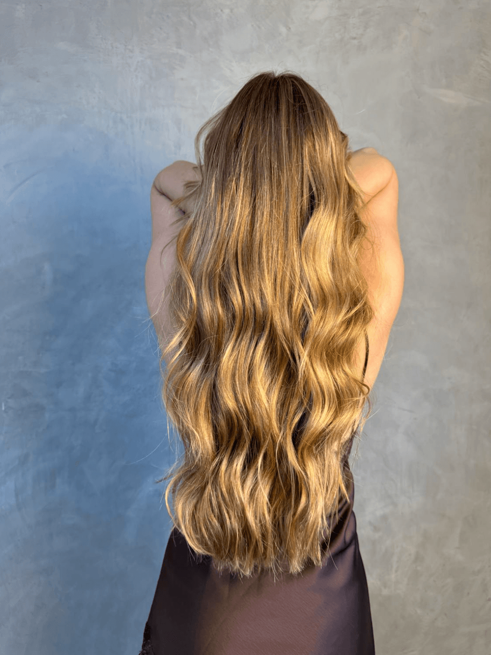 Woman with long, wavy blonde hair facing away against a gray background.