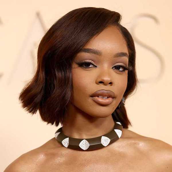 Woman with short hair and bold necklace, wearing elegant makeup against a light background.