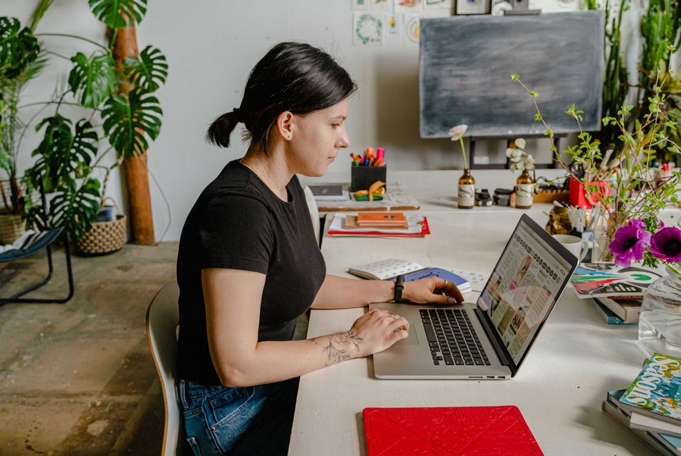 Woman working on a laptop at a plant-filled desk, surrounded by art supplies.