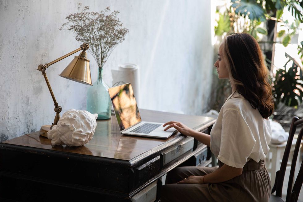 Woman working on a laptop at a wooden desk with plants and a brass lamp nearby.
