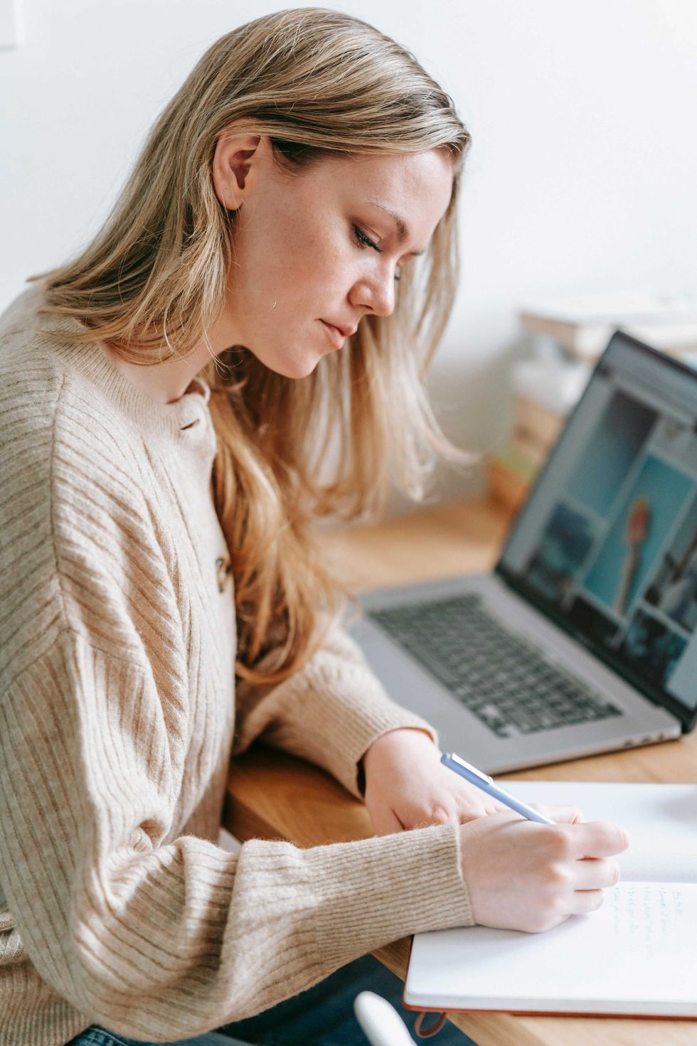 Woman writing in a notebook beside a laptop on a desk.