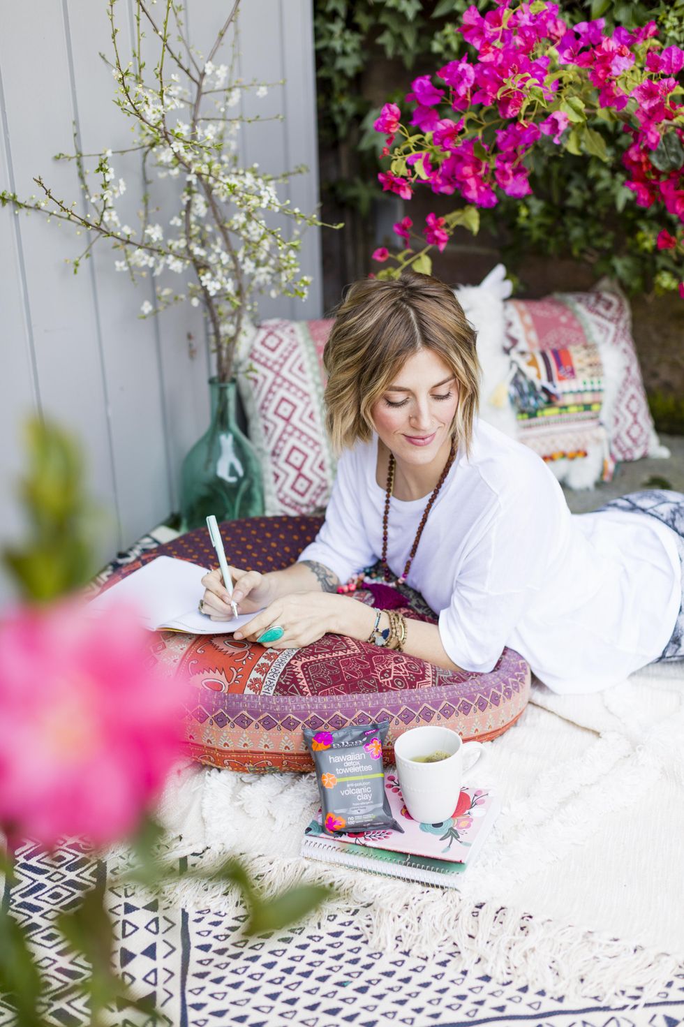 Woman writing while lounging outside, surrounded by flowers and cushions.