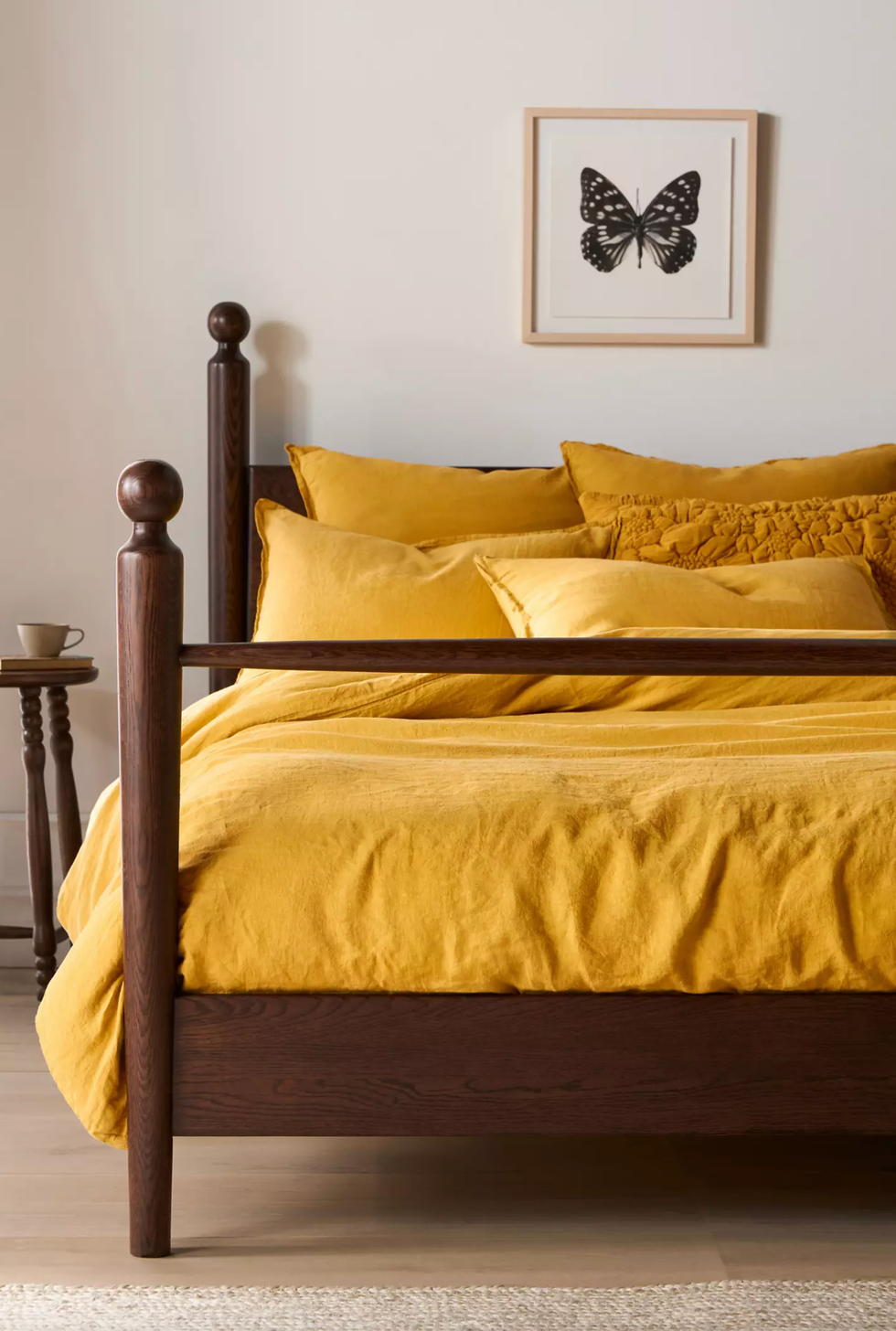 Wooden bed with mustard bedding, butterfly art, side table with mug.
