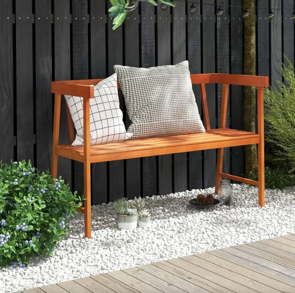Wooden bench with cushions on gravel, black fence, and plants nearby.