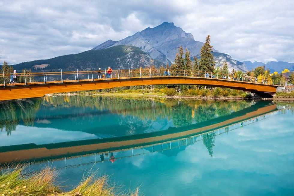 Wooden bridge over a turquoise river, with mountains and sky reflected in the water.