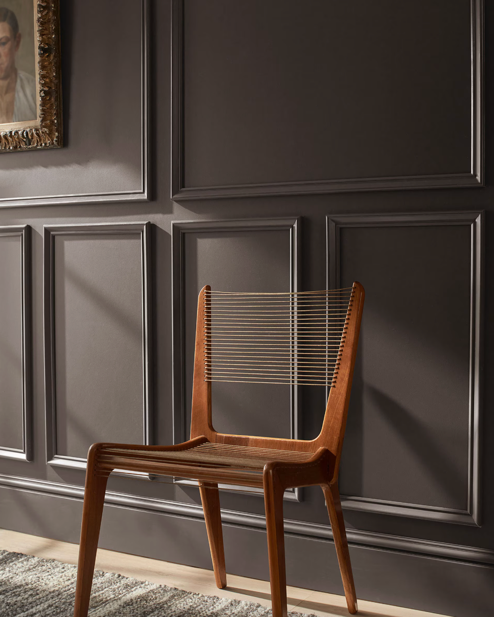 Wooden chair with slatted back against a paneled dark wall, near artwork.