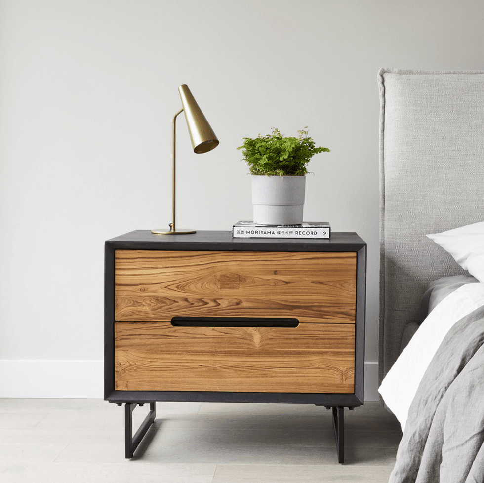 Wooden nightstand with lamp, plant, and book beside a bed with light gray headboard.